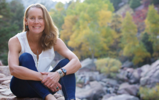 A white woman with blonde hair poses on a rock with fall foliage in the background