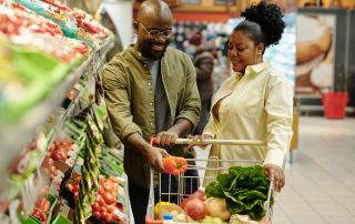 Happy man putting fresh pepper into shopping cart in supermarket with his wife